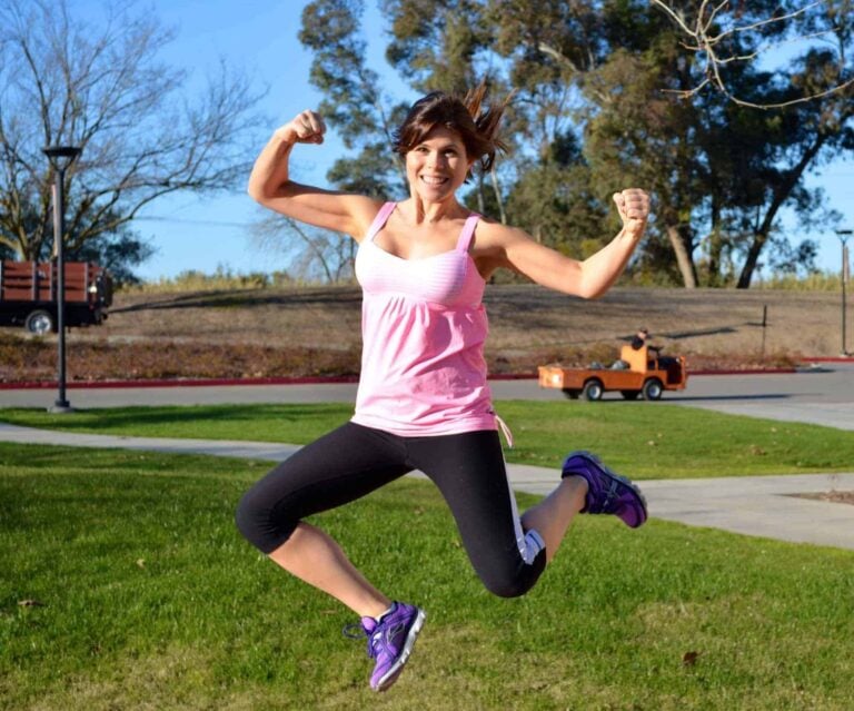 A Joyful Woman Wearing A Pink Tank Top And Black Leggings Is Leaping Into The Air With Her Fists Pumped In A Display Of Energy And Happiness, Set Against A Park Backdrop With Clear Blue Skies, Celebrating
