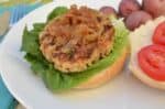 Grilled Turkey Burger Topped With Caramelized Onions And Spring Asparagus, Served On A Bun With Lettuce And Tomatoes, Alongside A Side Of Red Potatoes.