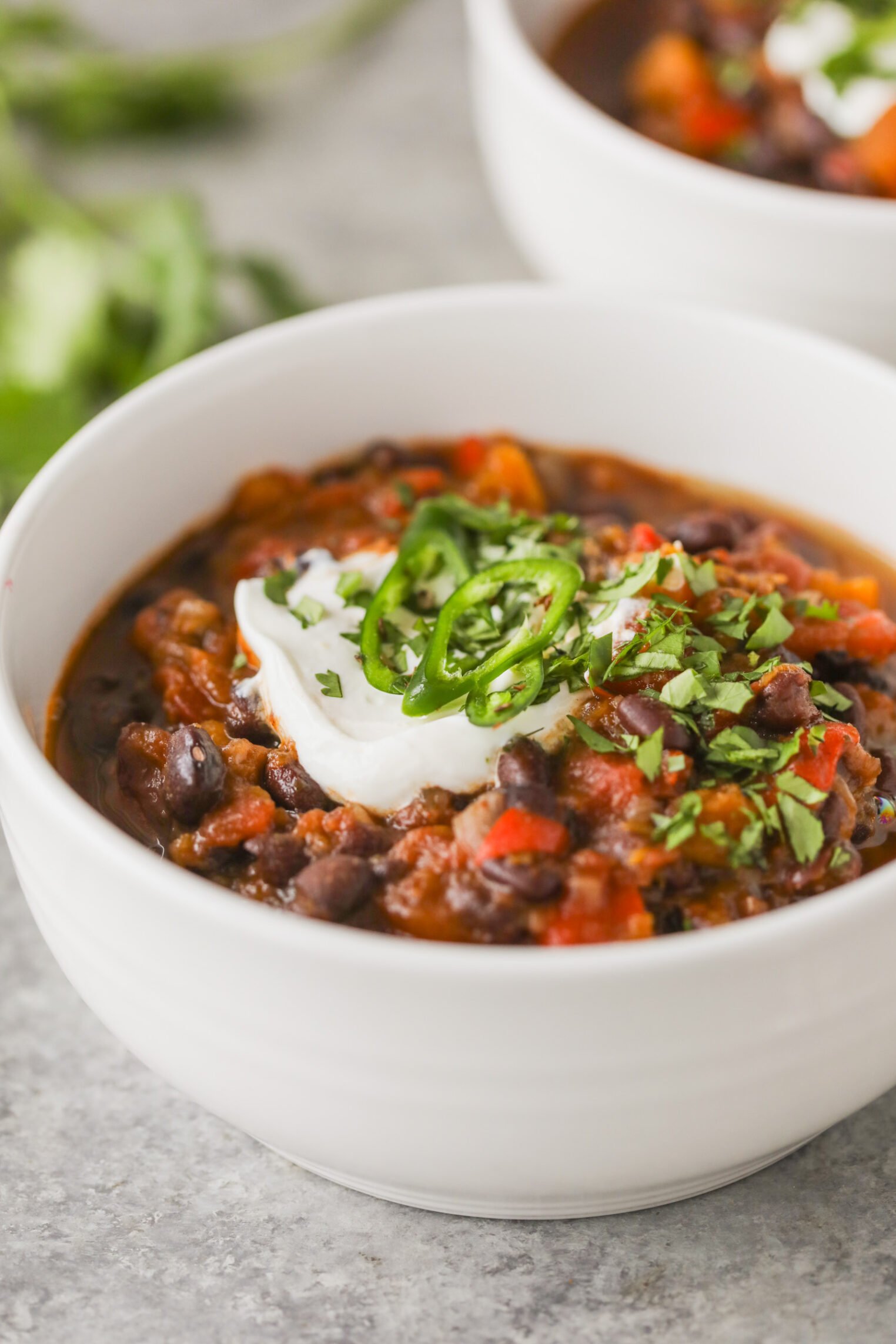 Slow Cooker Black Bean Chili with Butternut Squash 7 A White Bowl Filled With Slow Cooker Black Bean Chili, Topped With A Dollop Of Sour Cream, Sliced Jalapeños, And Chopped Cilantro. The Hearty Chili Features Black Beans, Tomatoes, And Peppers. Another Bowl Is Partially Visible In The Background.