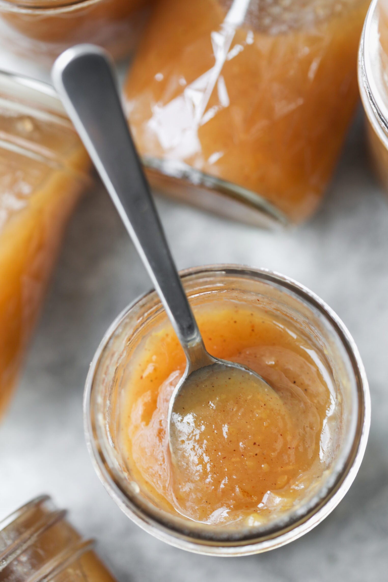 A Spoon In A Glass Jar Of Homemade Applesauce, Surrounded By Other Jars Filled With The Same Chunky, Light Brown Sauce Reminiscent Of Spiced Apple Butter.