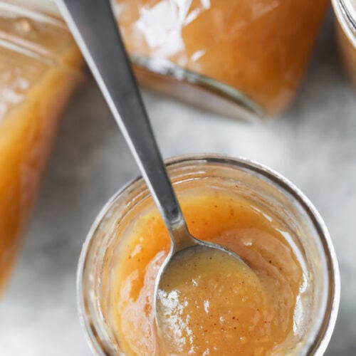 A Spoon In A Glass Jar Of Homemade Applesauce, Surrounded By Other Jars Filled With The Same Chunky, Light Brown Sauce Reminiscent Of Spiced Apple Butter.