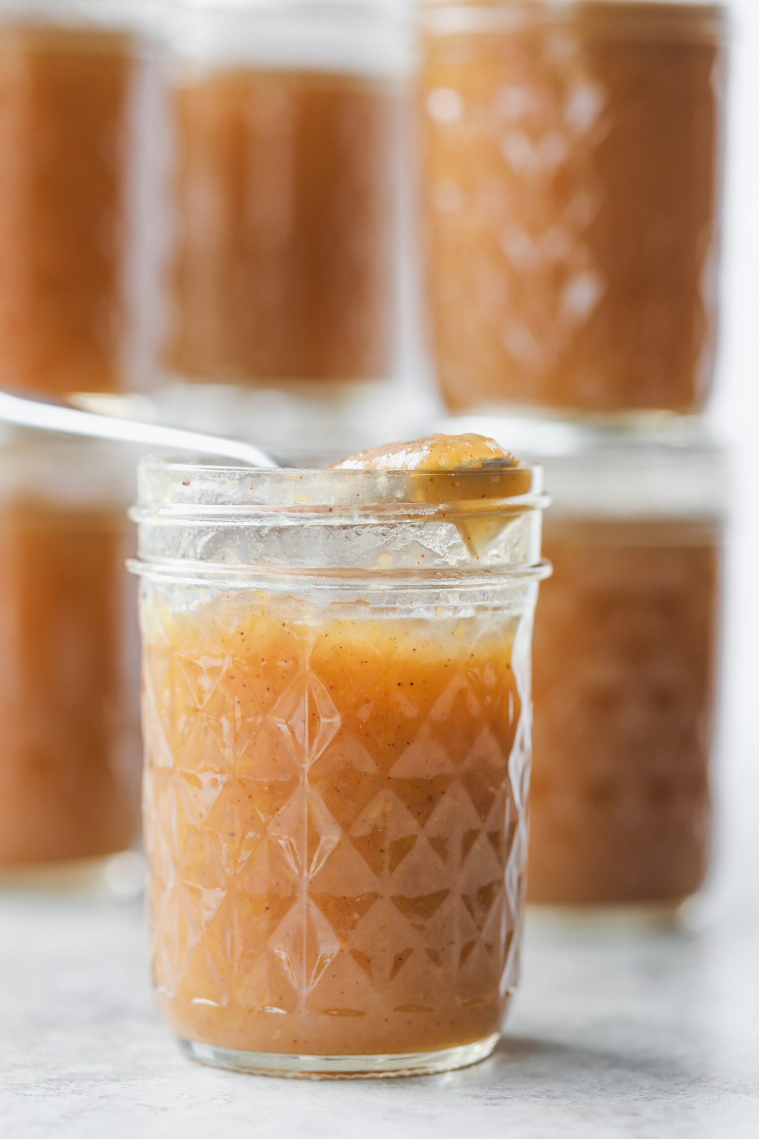 A Spoon Scoops Rich Apple Butter From A Glass Jar, With More Jars Of Homemade Apple Butter Blurred In The Background.