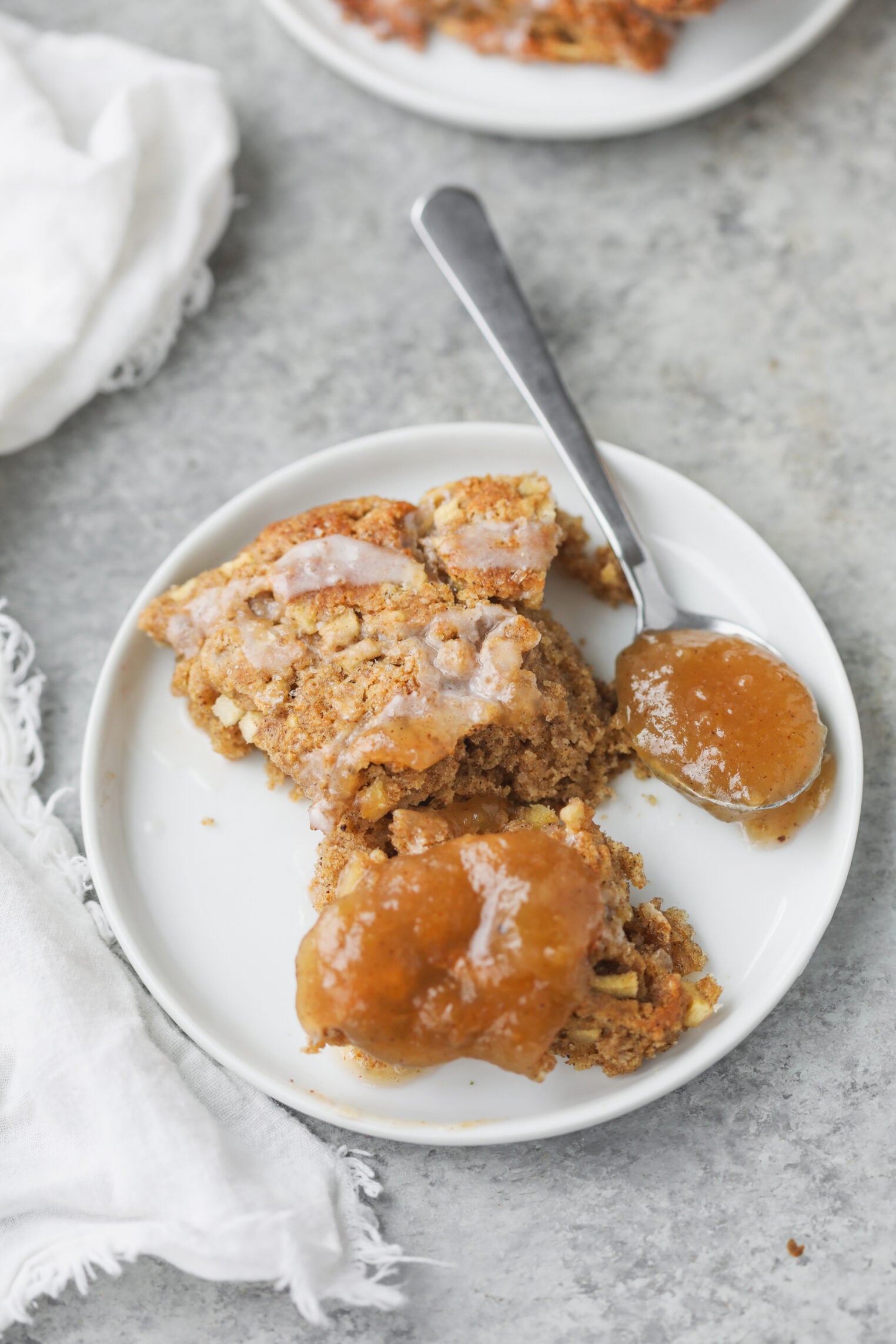 A White Plate With A Serving Of Apple Crumble Topped With Glaze, A Spoonful Of Rich Apple Butter On The Side, And A Silver Spoon. The Plate Is On A Gray Surface With A White Napkin Nearby.