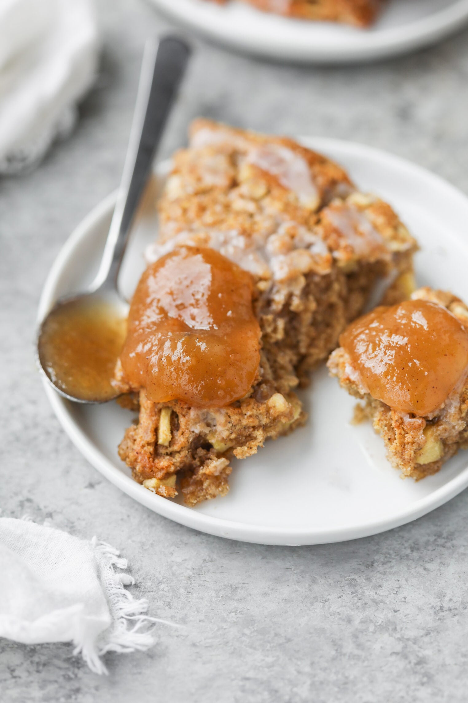 A White Plate With Slices Of Spiced Cake Topped With Thick Applesauce And A Dollop Of Rich Apple Butter, Alongside A Silver Spoon, On A Gray Surface With A White Napkin Nearby.