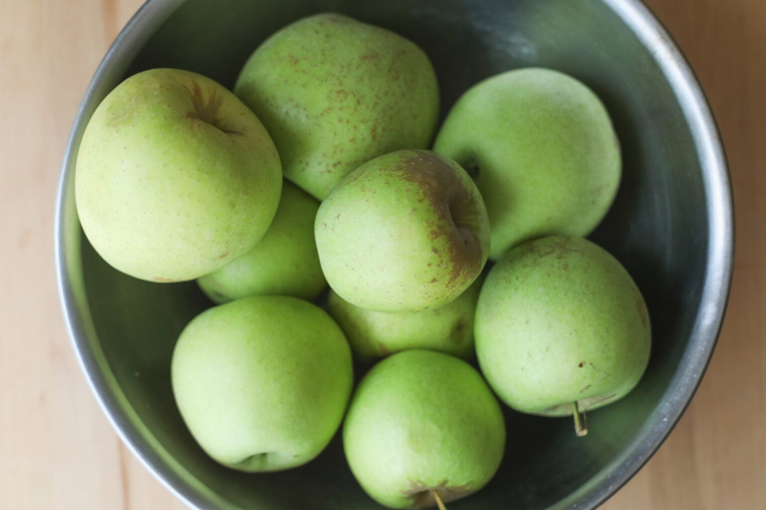 A Metal Bowl Filled With Several Light Green Apples, Perfect For Making Homemade Apple Butter, Sits On A Wooden Surface.