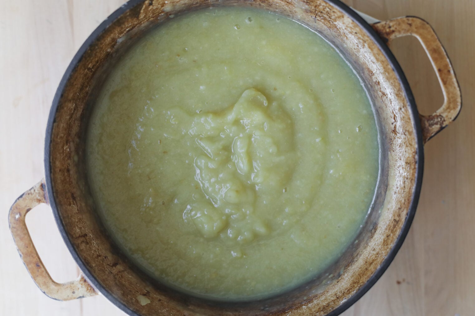 A Top View Of A Rustic Pot Containing Thick, Green Soup Or Puree, Likely Made From Blended Vegetables And Subtly Infused With Apple Butter, Sitting On A Light Wooden Surface.