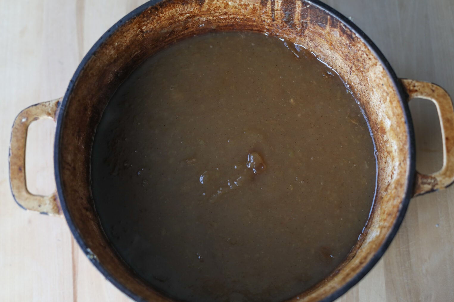 A Large, Rustic Pot Filled With Thick, Brown Apple Butter Sits On A Light Wooden Surface. The Pot Has Visible Stains And Wear Around The Edges.