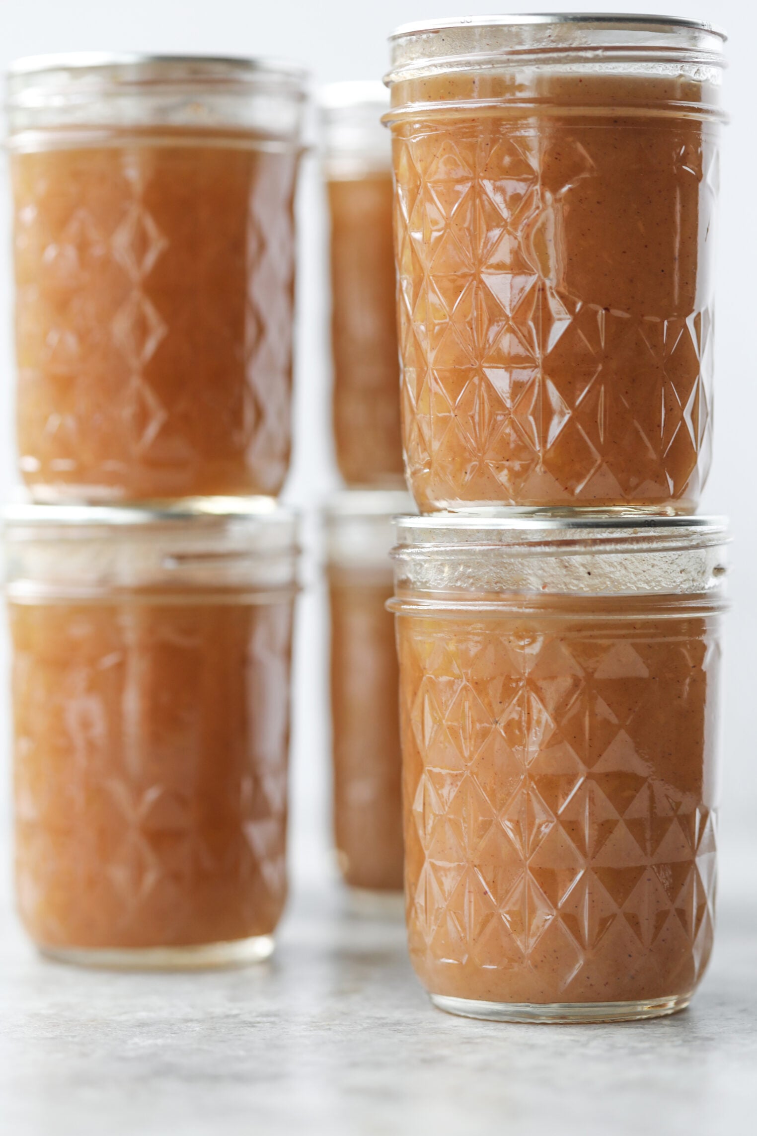Six Glass Jars Filled With Light Brown Applesauce And Apple Butter Are Stacked In Two Columns On A Light Gray Surface, With A Neutral Background.