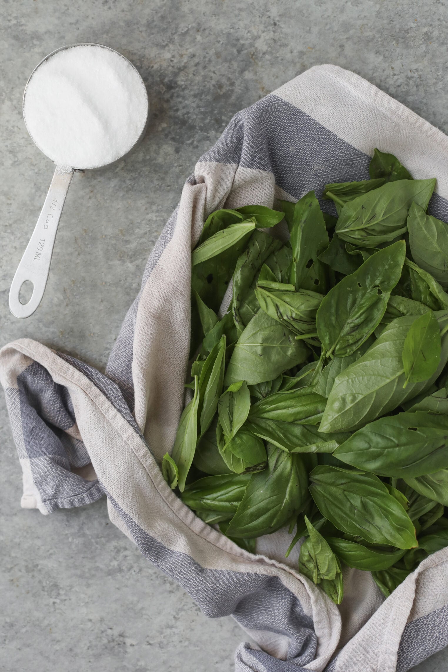 Fresh Basil Leaves On A Striped Towel Next To A Metal Measuring Cup Filled With Granulated Sugar, Placed On A Gray Stone Surface.