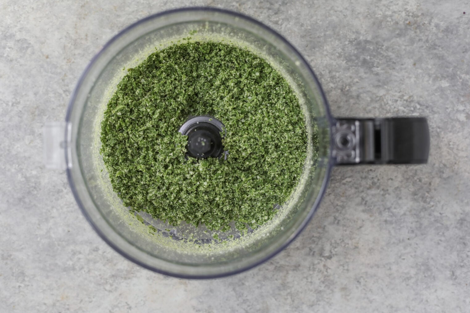A Food Processor Bowl Filled With Finely Chopped Green Herbs Sits On A Gray Countertop, Seen From Above.