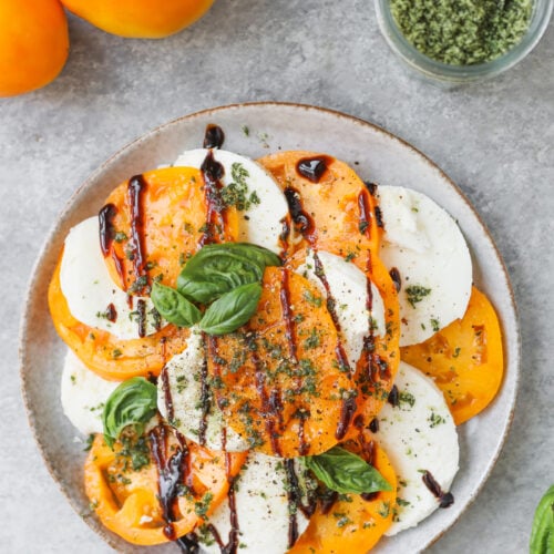 A Plate Of Yellow Tomato And Mozzarella Slices Topped With Basil Leaves, Pesto, And A Drizzle Of Balsamic Glaze Sits On A Gray Surface With Whole Tomatoes And A Jar Of Pesto Nearby.