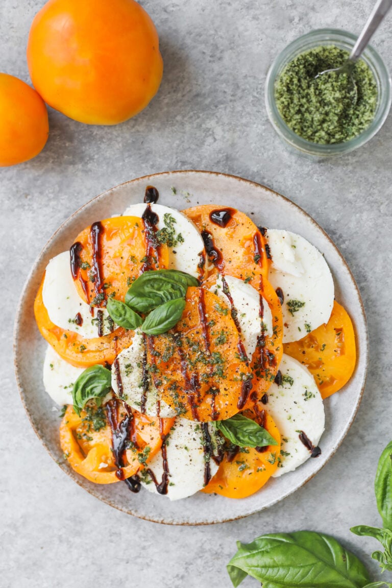 A Plate Of Yellow Tomato And Mozzarella Slices Topped With Basil Leaves, Pesto, And A Drizzle Of Balsamic Glaze Sits On A Gray Surface With Whole Tomatoes And A Jar Of Pesto Nearby.