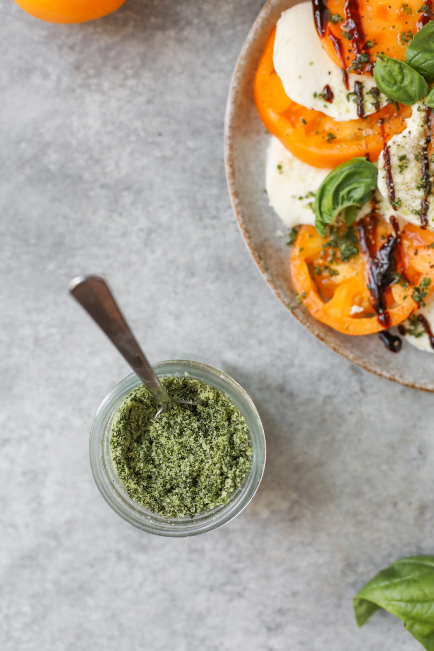 A Jar Of Green Herb Salt With A Spoon Sits Next To A Plate Of Caprese Salad With Yellow Tomatoes, Mozzarella, Basil, And A Drizzle Of Balsamic Glaze On A Gray Surface.