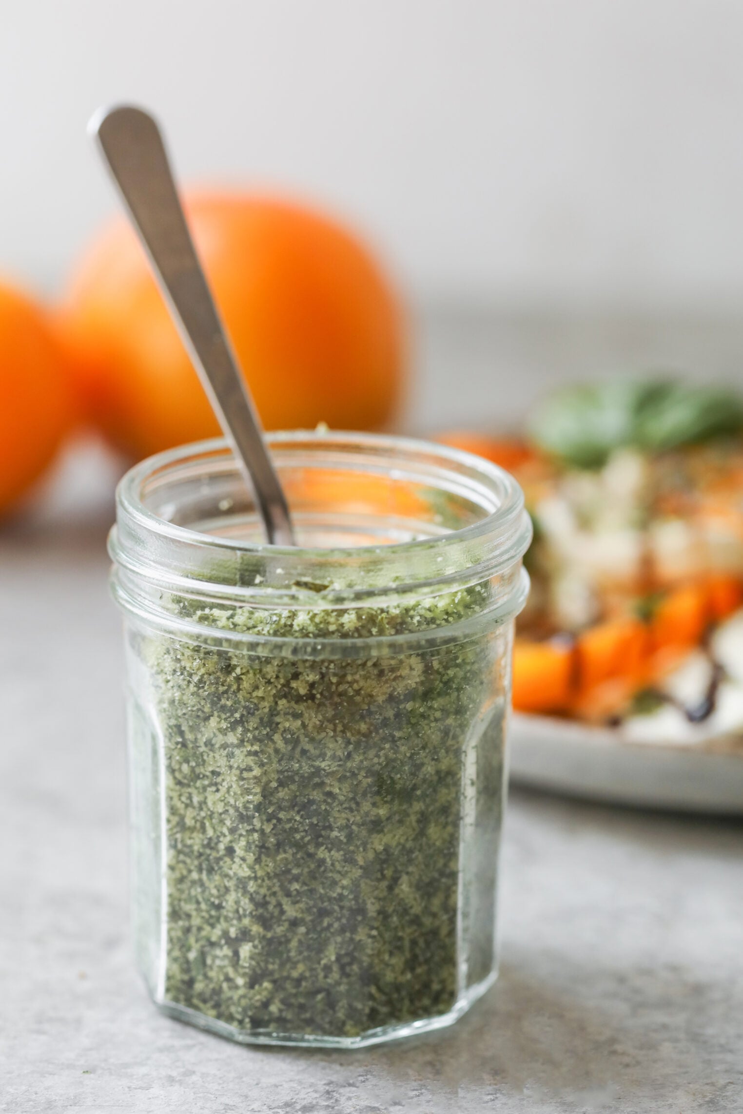 A Glass Jar Filled With Green Pesto And A Spoon Inside, With Blurred Oranges And A Plate Of Food In The Background.