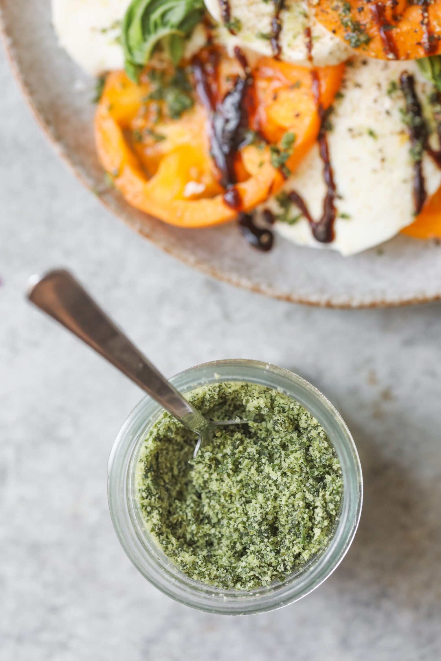 A Small Glass Jar Of Green Herb Salt With A Spoon Inside, Placed On A Gray Surface. In The Background, There Is A Plate Of Sliced Tomatoes, Mozzarella, Fresh Basil, And A Drizzle Of Balsamic Glaze.