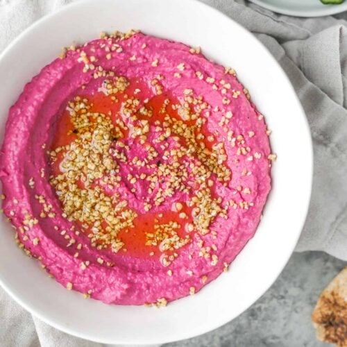 A Bowl Of Vibrant Pink Beet Hummus Is Artfully Garnished With Sesame Seeds And A Drizzle Of Olive Oil. Placed Elegantly On A Gray Surface, It'S Surrounded By Crisp Cucumber Slices And Warm Pita Bread, Inviting You To Savor The Delightful Flavors.