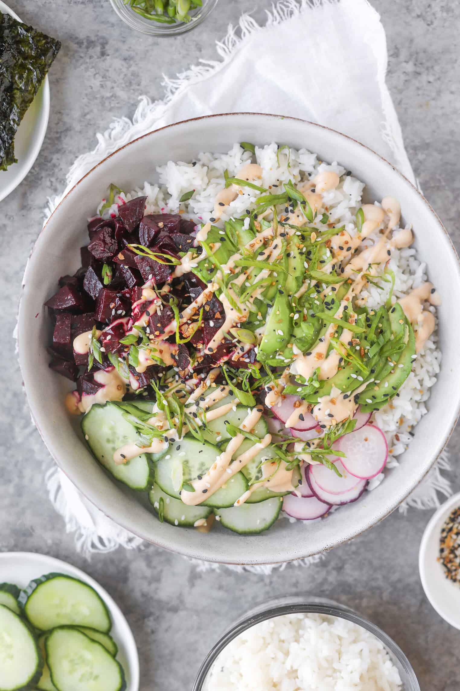 Beet Poke Bowls 10 A Bowl Of White Rice Topped With Sliced Cucumbers, Radishes, Avocado, Diced Beets, Green Onions, Sesame Seeds, And A Creamy Orange Sauce—This Vibrant Beet Poke Bowl Sits On A White Napkin With Other Small Dishes Nearby.