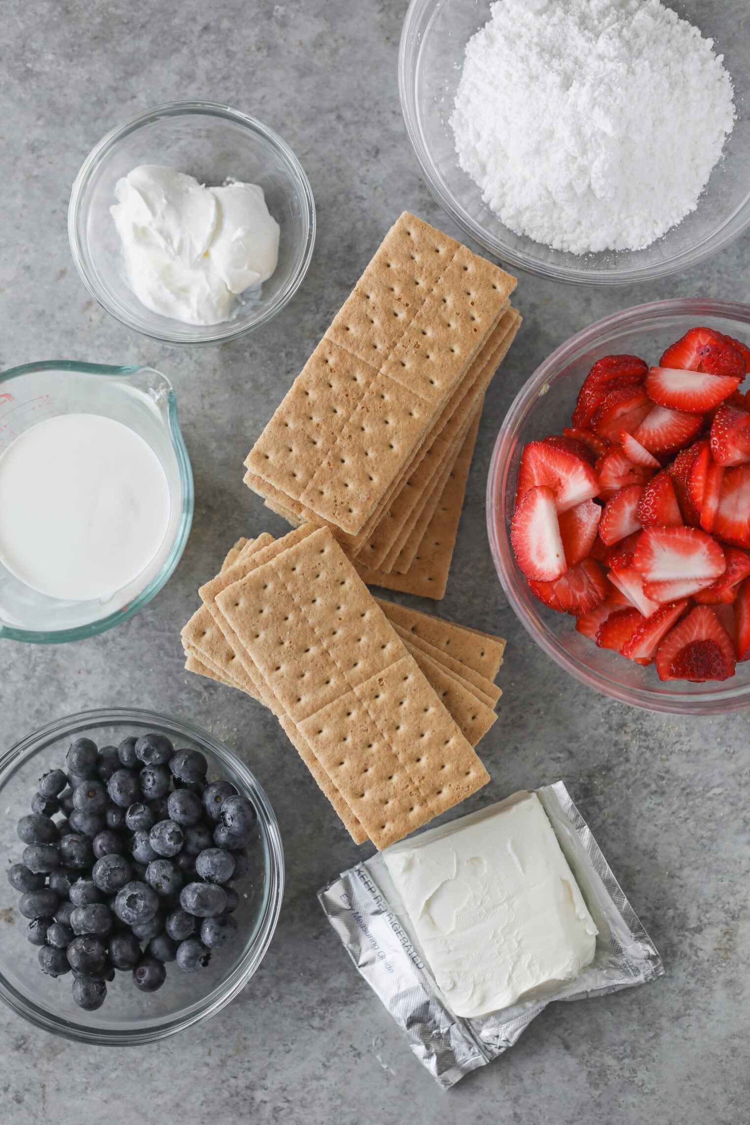 Berry Icebox Cake 2 Bowls With Powdered Sugar, Whipped Cream, Yogurt, Blueberries, Sliced Strawberries, Cream Cheese, And A Stack Of Graham Crackers Arranged On A Gray Surface—Perfect Ingredients For Assembling A Classic Berry Icebox Cake.