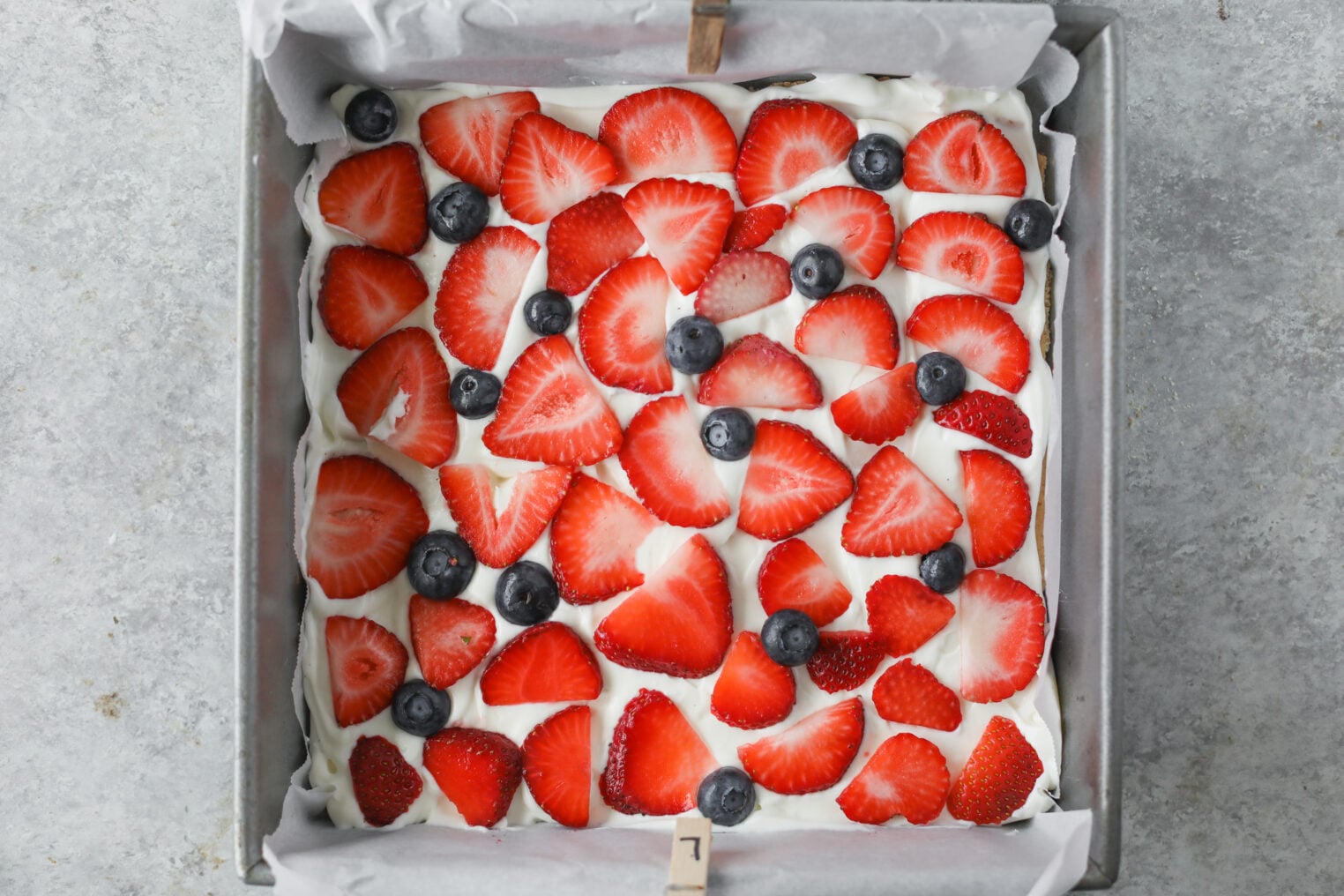 Berry Icebox Cake 8 A Square Berry Icebox Cake Topped With A Layer Of Whipped Cream, Sliced Strawberries, And Whole Blueberries, In A Parchment-Lined Baking Pan On A Gray Surface.