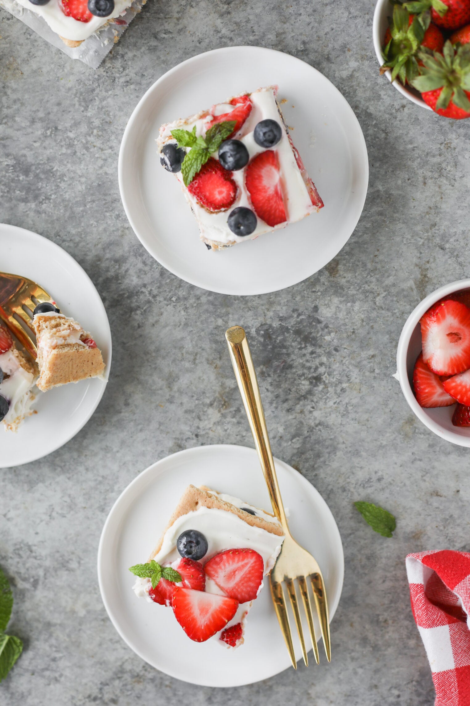 Berry Icebox Cake 10 Two Plates With Berry Icebox Cake Topped With Strawberries, Blueberries, And Mint Sit On A Gray Surface. A Gold Fork Rests On One Plate. Nearby, Bowls Of Fresh Strawberries And A Red-White Napkin Are Visible.