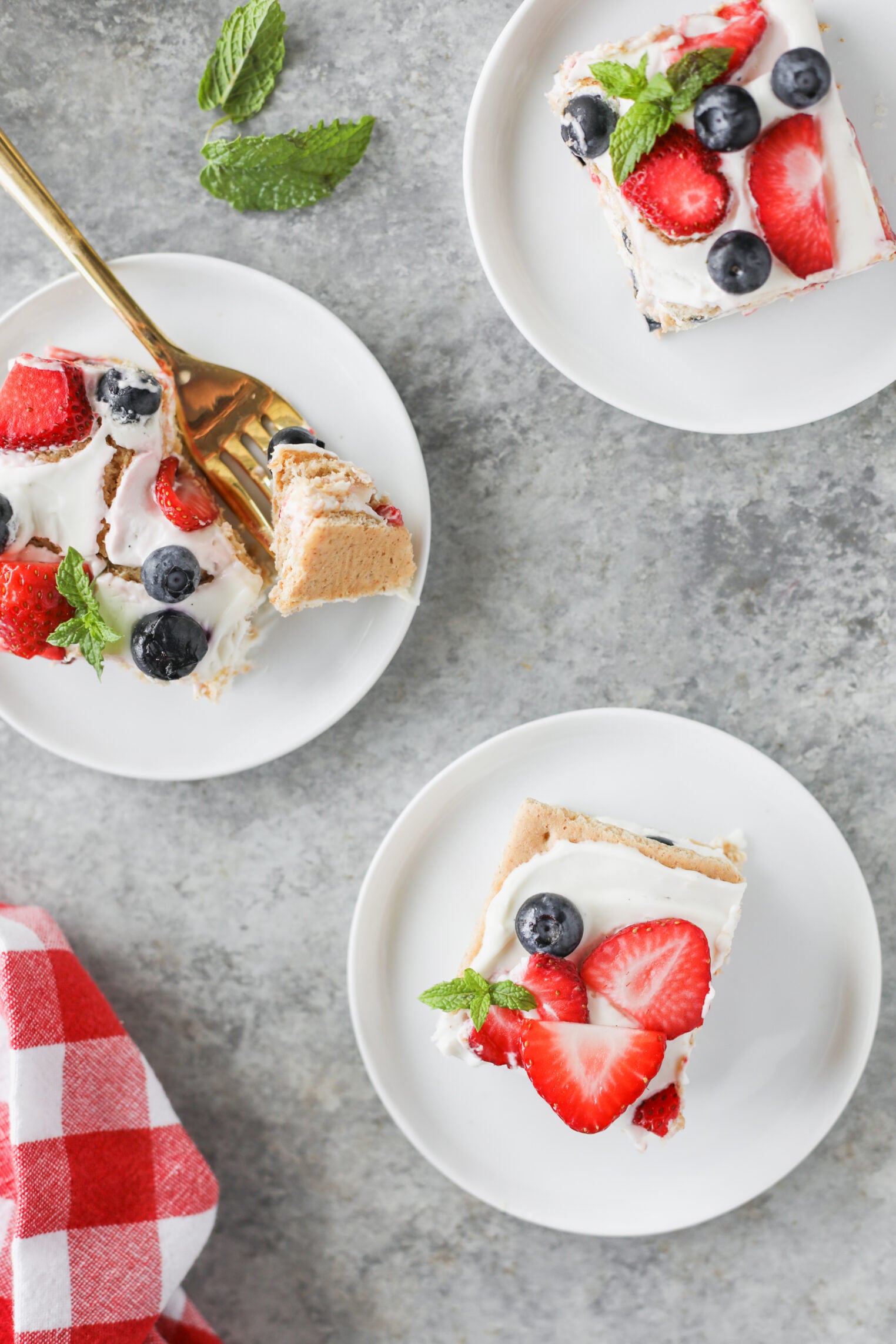 Berry Icebox Cake 3 Three Berry Icebox Cake Dessert Bars Topped With Whipped Cream, Strawberries, And Blueberries Are On Small White Plates, With A Gold Fork Holding A Bite. A Red-Checkered Cloth And Mint Leaves Are Nearby On A Gray Surface.