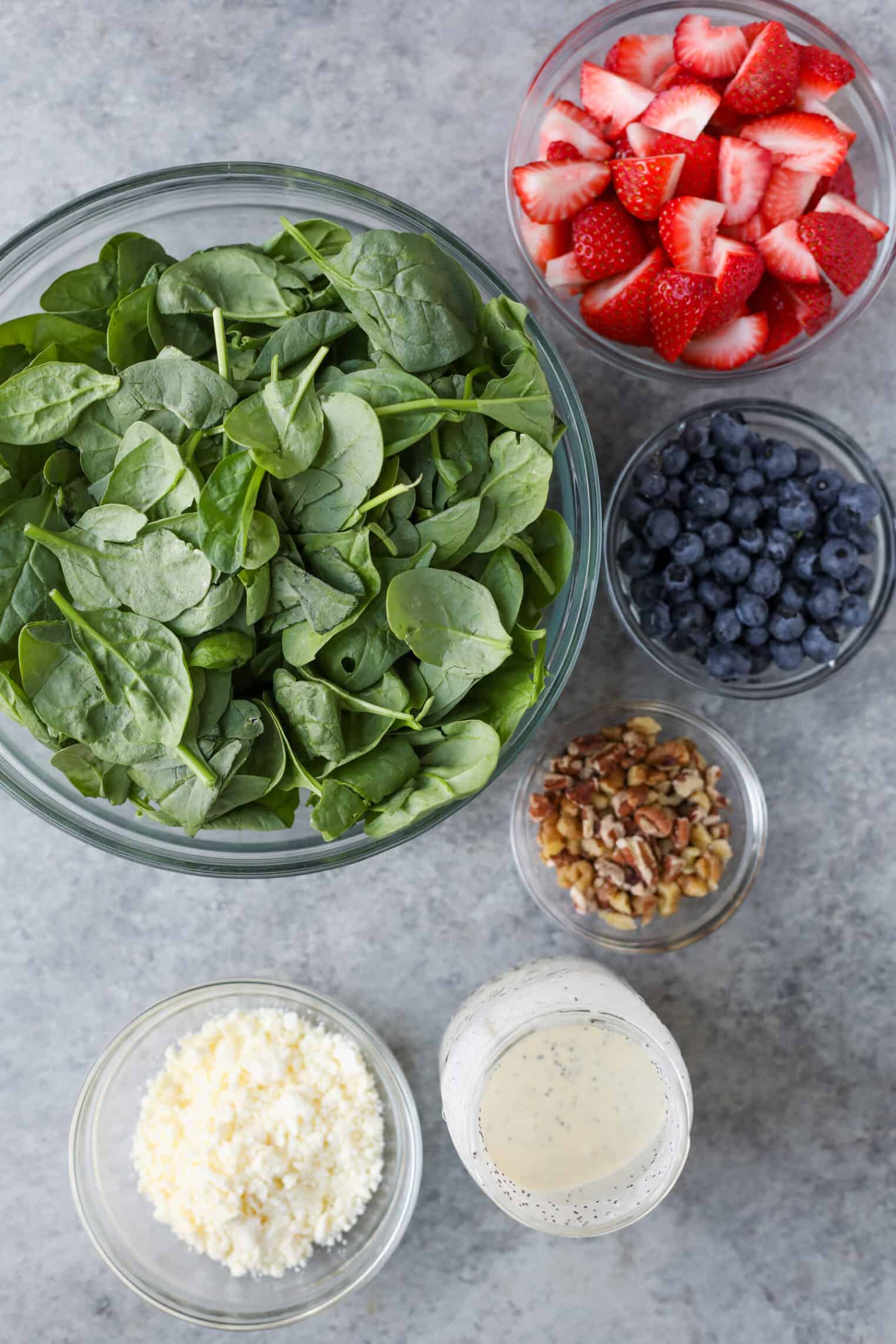 Strawberry Poppyseed Salad 2 A Flat Lay Of Salad Ingredients: A Bowl Of Fresh Spinach, Sliced Strawberry, Blueberries, Chopped Pecans, Crumbled Cheese, And A Glass Of Creamy Poppy Seed Salad Dressing On A Gray Surface.