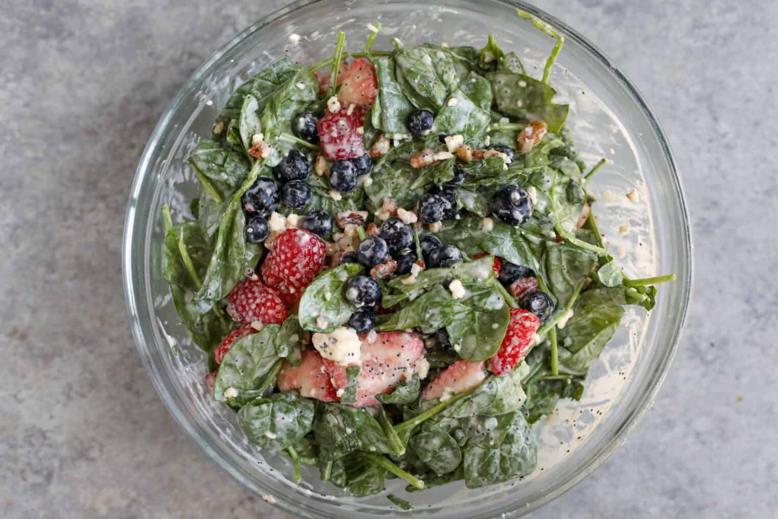 Strawberry Poppyseed Salad 4 A Glass Bowl Filled With Strawberry Poppy Seed Salad—Spinach Topped With Strawberries, Blueberries, Crumbled Cheese, Chopped Nuts, And Creamy Dressing—Sits On A Gray Surface.
