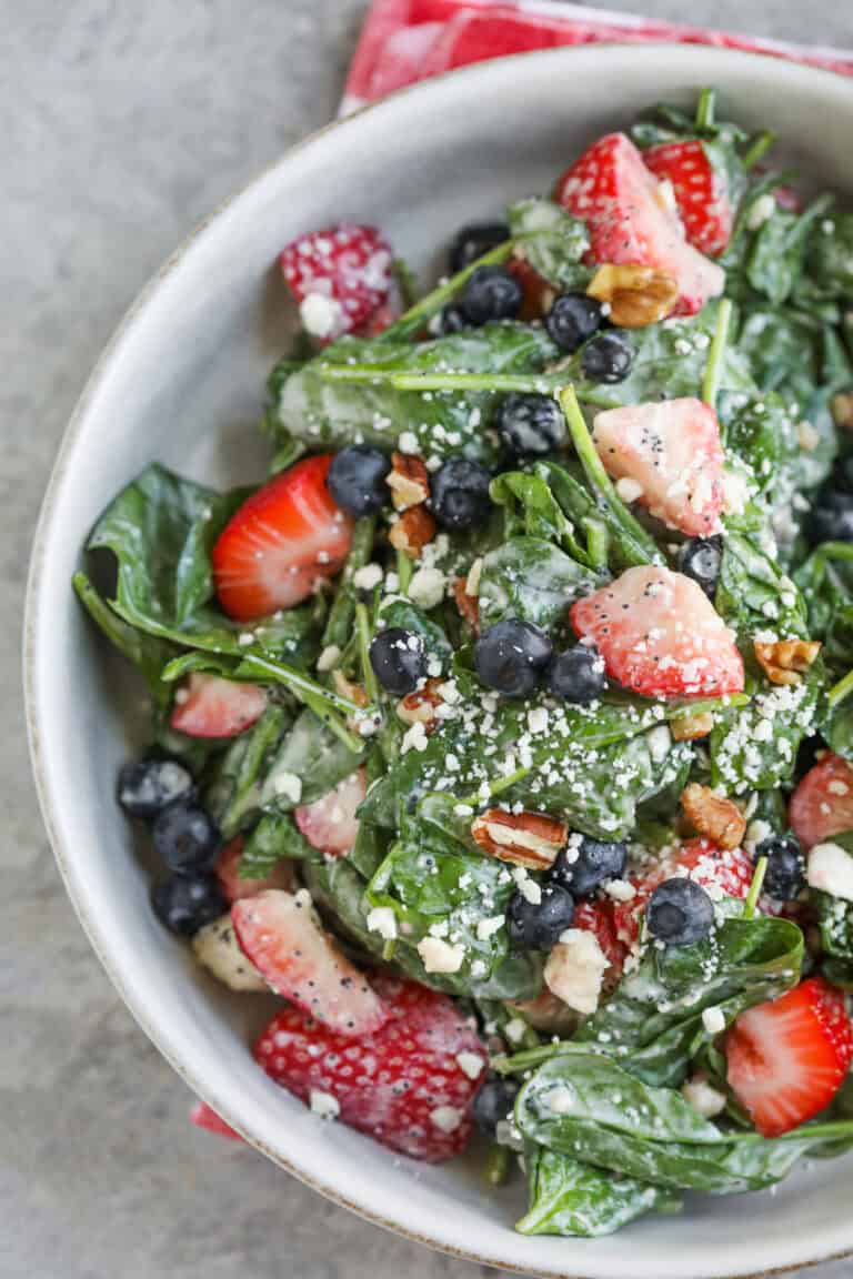 A Bowl Of Fresh Spinach Salad With Strawberries, Blueberries, Pecans, Crumbled Cheese, And A Creamy Poppy Seed Dressing, Set Against A Light Background With A Red Napkin Partially Visible.