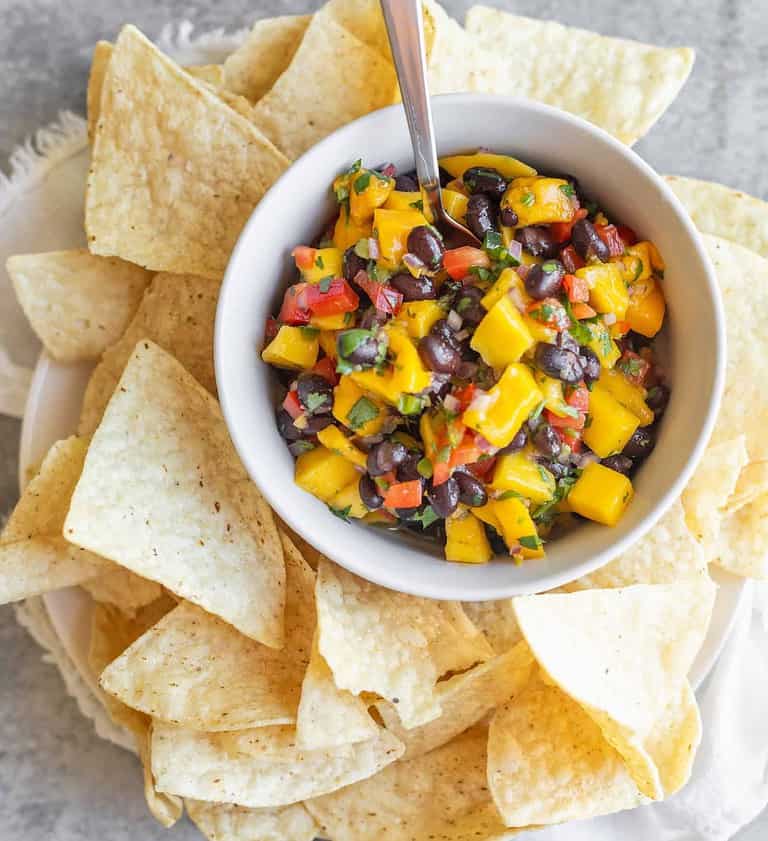 A Bowl Of Black Bean And Mango Salsa With Diced Red Peppers And Herbs Sits On A Plate, Surrounded By Tortilla Chips. This Vibrant Dish Makes For One Of The Best Healthy Appetizers. A Spoon Is In The Bowl, Set Against A Light Gray Textured Background.