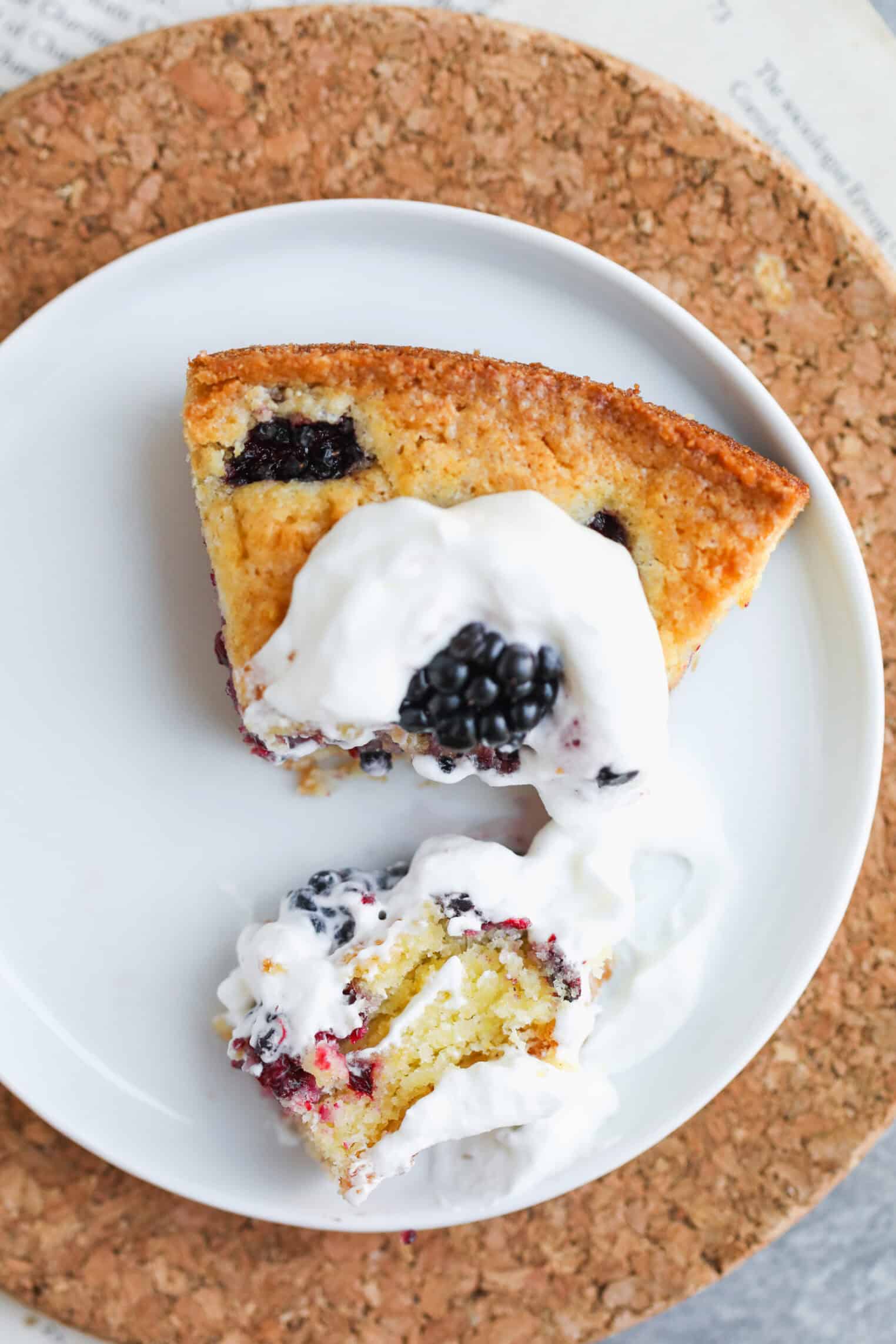 Blackberry Lemon Polenta Cake 11 A Slice Of Berry-Studded Polenta Cake Topped With Whipped Cream Sits On A White Plate, With One Bite Cut Out And Some Whipped Cream And A Blackberry On The Side. The Plate Rests On A Round Cork Mat.