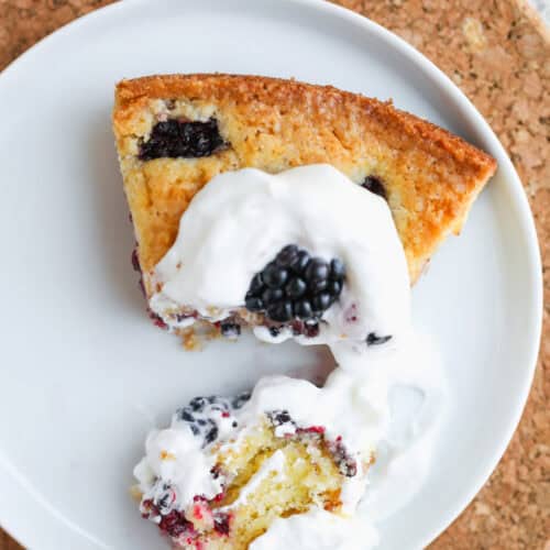 Blackberry Lemon Polenta Cake 13 A Slice Of Berry-Studded Polenta Cake Topped With Whipped Cream Sits On A White Plate, With One Bite Cut Out And Some Whipped Cream And A Blackberry On The Side. The Plate Rests On A Round Cork Mat.