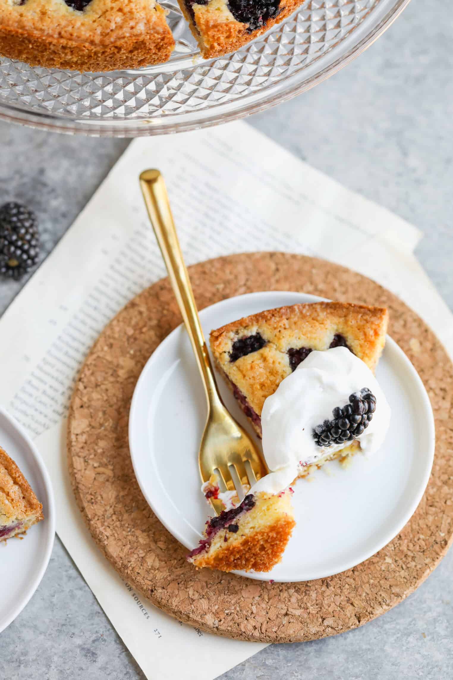 Blackberry Lemon Polenta Cake 1 A Slice Of Blackberry Polenta Cake Topped With Whipped Cream Is Served On A White Plate With A Gold Fork, Placed On A Cork Mat. More Cake Is Visible On A Glass Stand In The Background, With A Few Papers Beneath The Plate.