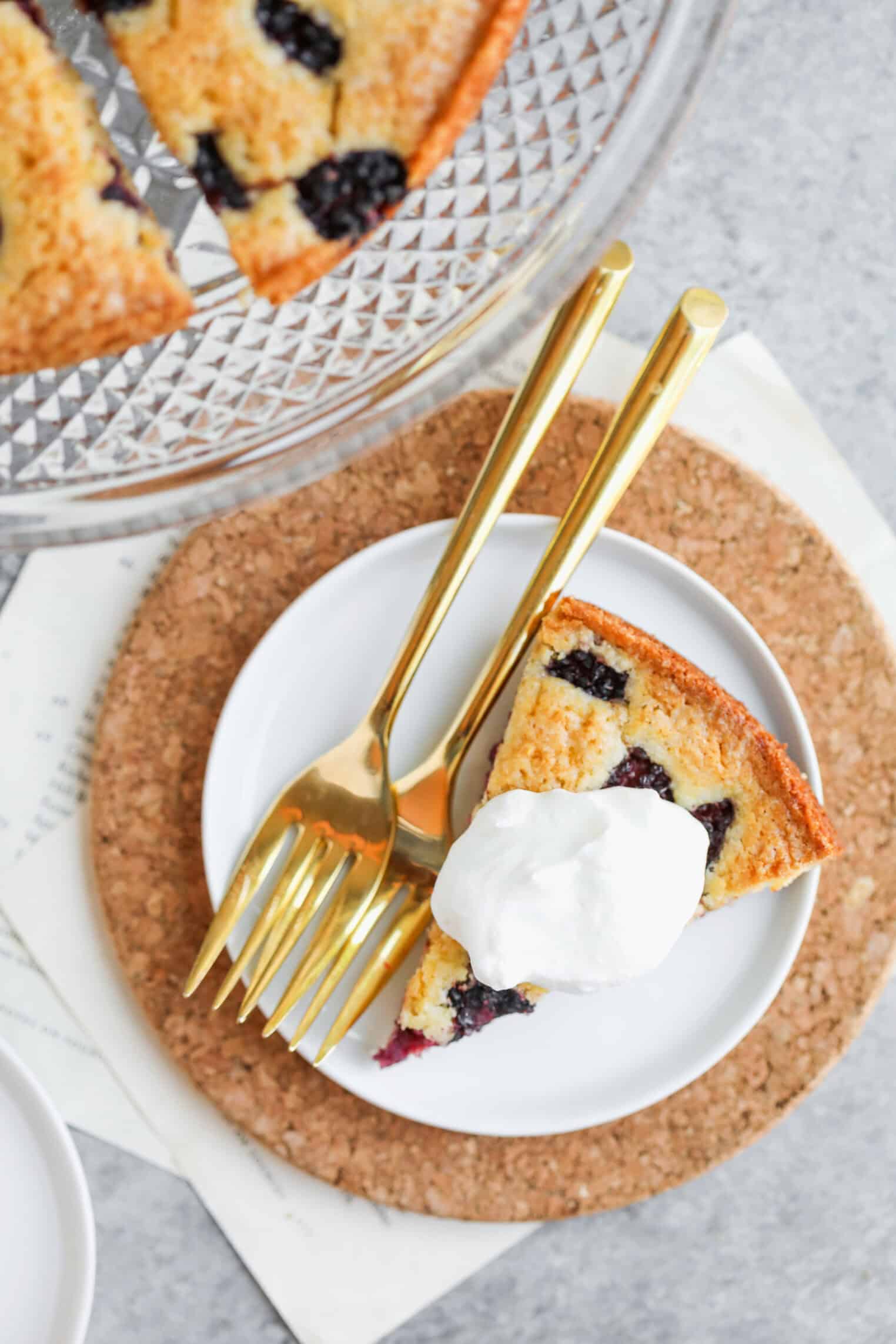 Blackberry Lemon Polenta Cake 3 A Slice Of Berry Lemon Polenta Cake Topped With Whipped Cream Sits On A White Plate With Two Gold Forks, Placed On A Cork Mat. A Cake Stand With More Cake Is Partially Visible Above.