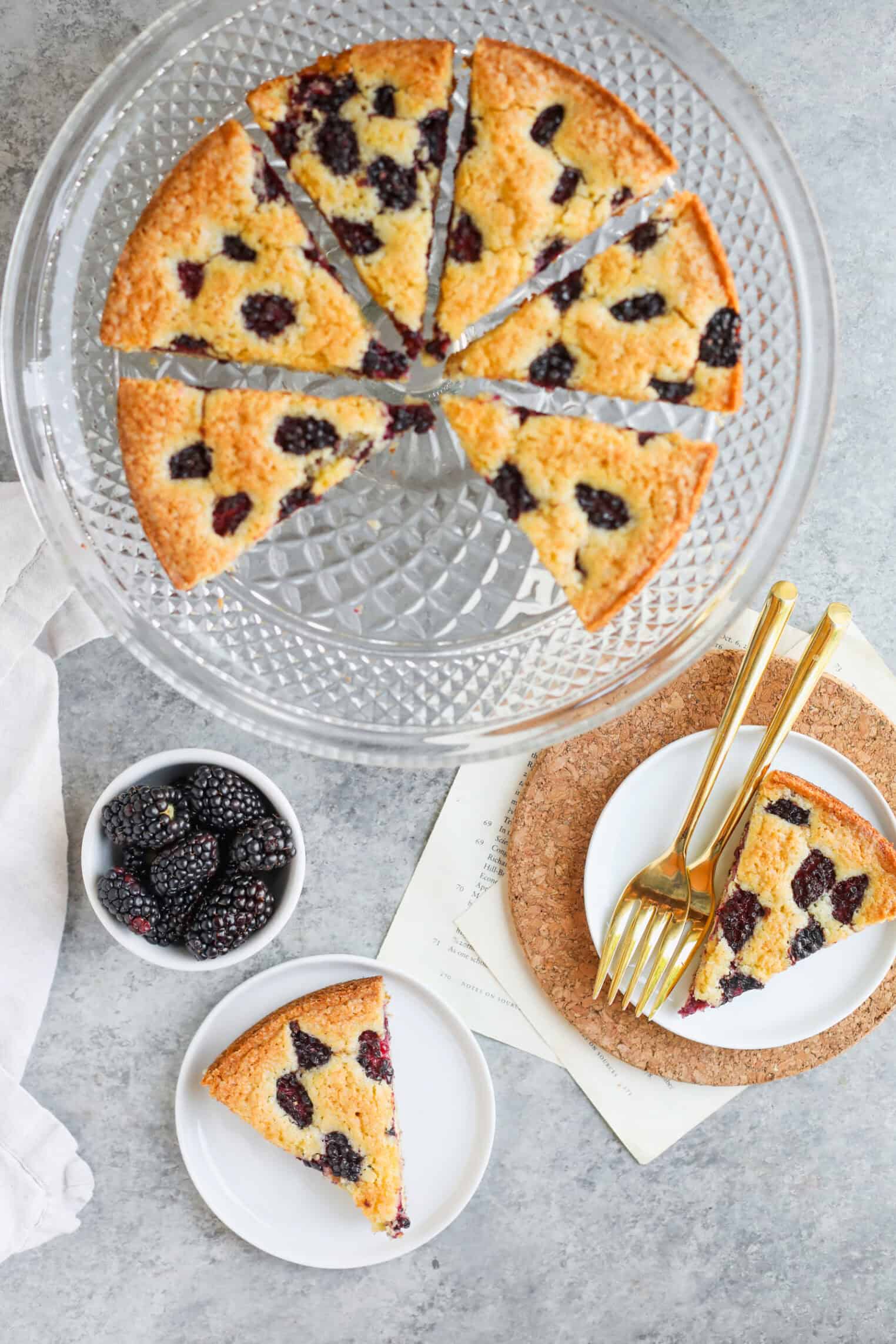 Blackberry Lemon Polenta Cake 2 A Sliced Blackberry Polenta Cake On A Glass Cake Stand, With One Lemon-Infused Slice Served On A Small White Plate And A Bowl Of Fresh Blackberries Nearby. Gold Forks Rest On Another Plate, All Set On A Light Gray Surface.