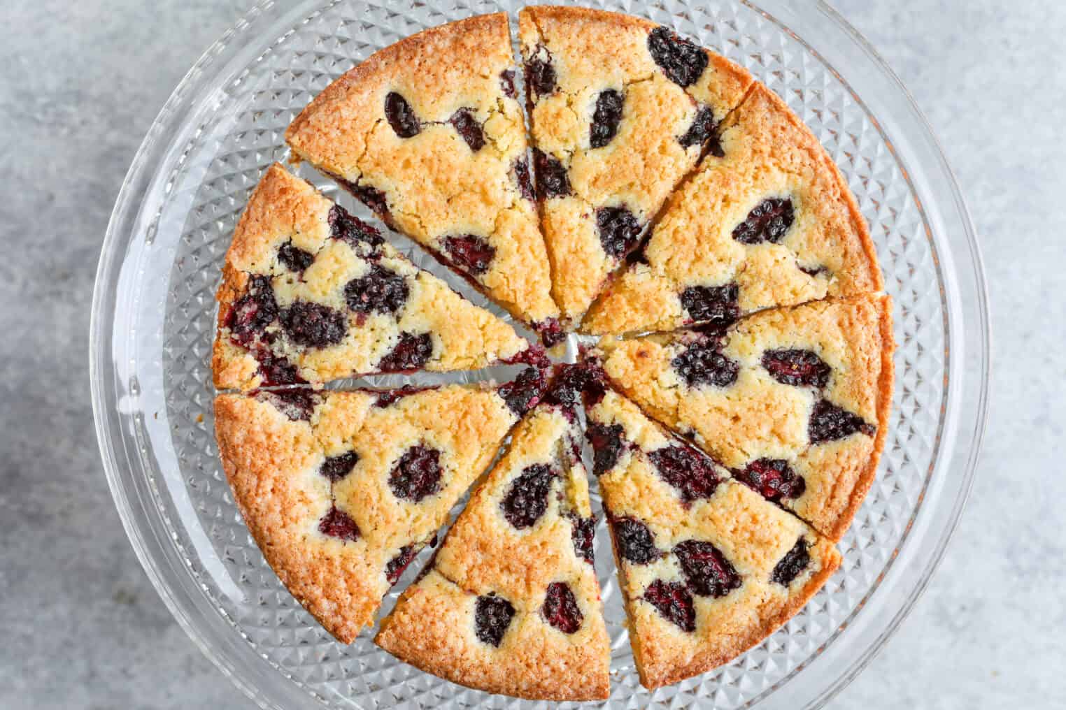 Blackberry Lemon Polenta Cake 10 A Round Blackberry-Studded Polenta Cake, Sliced Into Eight Pieces, Sits On A Clear Glass Cake Stand With A Textured Pattern. The Cake Has A Golden-Brown Crust And Visible Dark Berries Throughout.