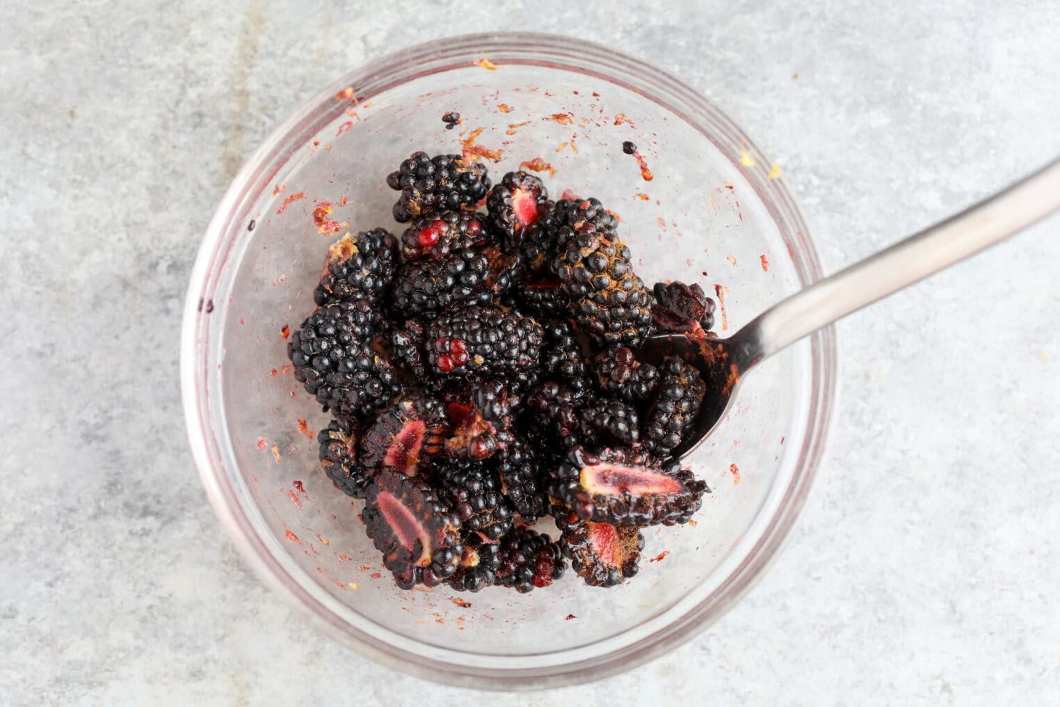 Blackberry Lemon Polenta Cake 7 A Clear Glass Bowl Filled With Blackberries And A Few Sliced Strawberries, Some Of The Fruit Slightly Mashed, With A Metal Spoon Resting Inside, On A Light Gray Textured Surface—Perfect As A Topping For Lemon Polenta Cake.