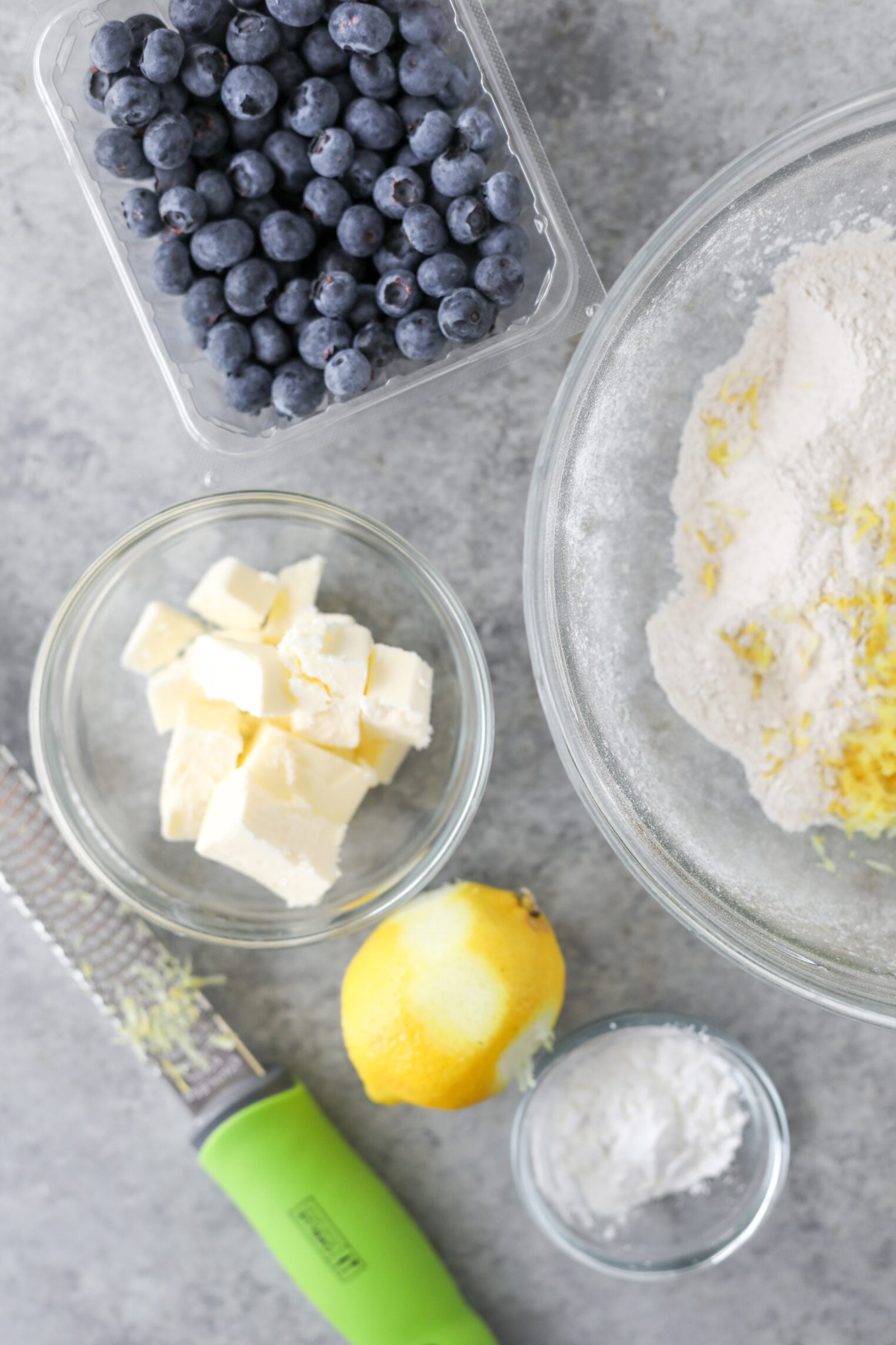 Overhead View Of Baking Ingredients For A Blueberry Galette: A Bowl Of Flour With Lemon Zest, A Container Of Blueberries, A Small Bowl Of Cubed Butter, A Whole Lemon, A Grater, And A Small Bowl Of White Powder, Likely Baking Powder.