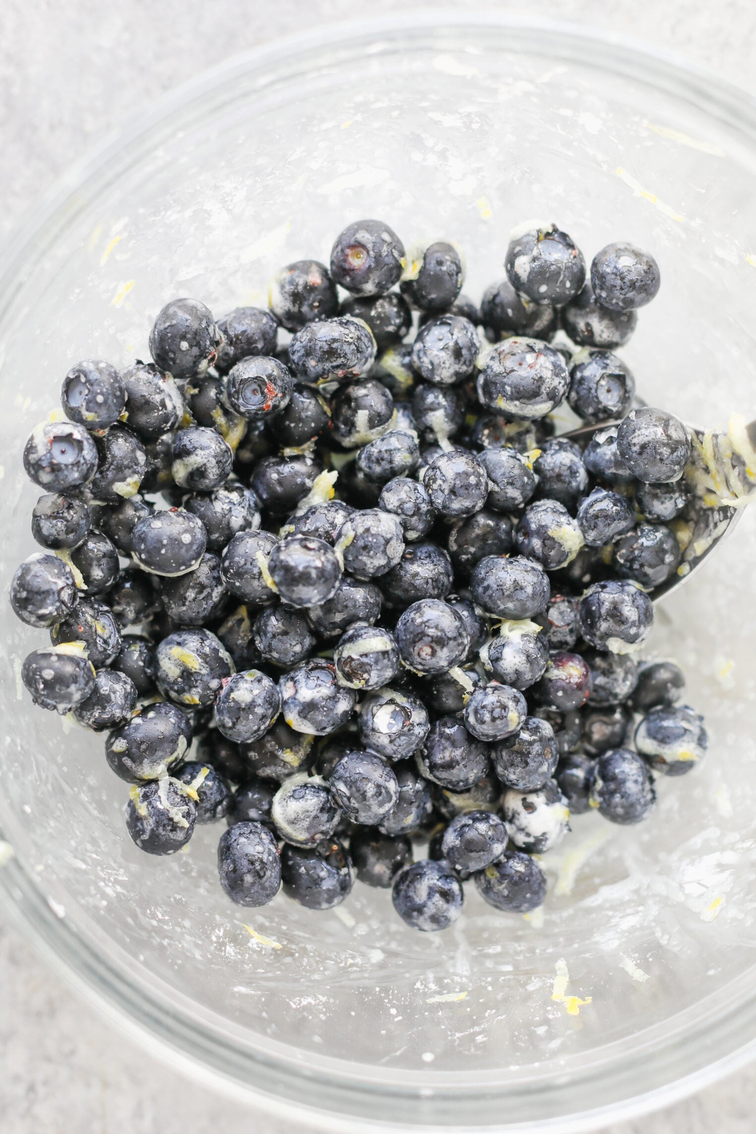 A Glass Bowl Filled With Fresh Blueberries Coated In A Light Mixture Of Flour And Lemon Zest, Just As You Would Prepare For A Homemade Blueberry Galette, With A Metal Spoon Partially Visible On The Right Side.