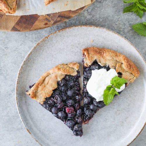 Two Slices Of Blueberry Galette Sit Invitingly On A Plate, One Crowned With A Dollop Of Whipped Cream And A Fresh Mint Sprig. The Plate Rests On A Gray Surface, Surrounded By Extra Blueberry Galette And A Bunch Of Mint Nearby.