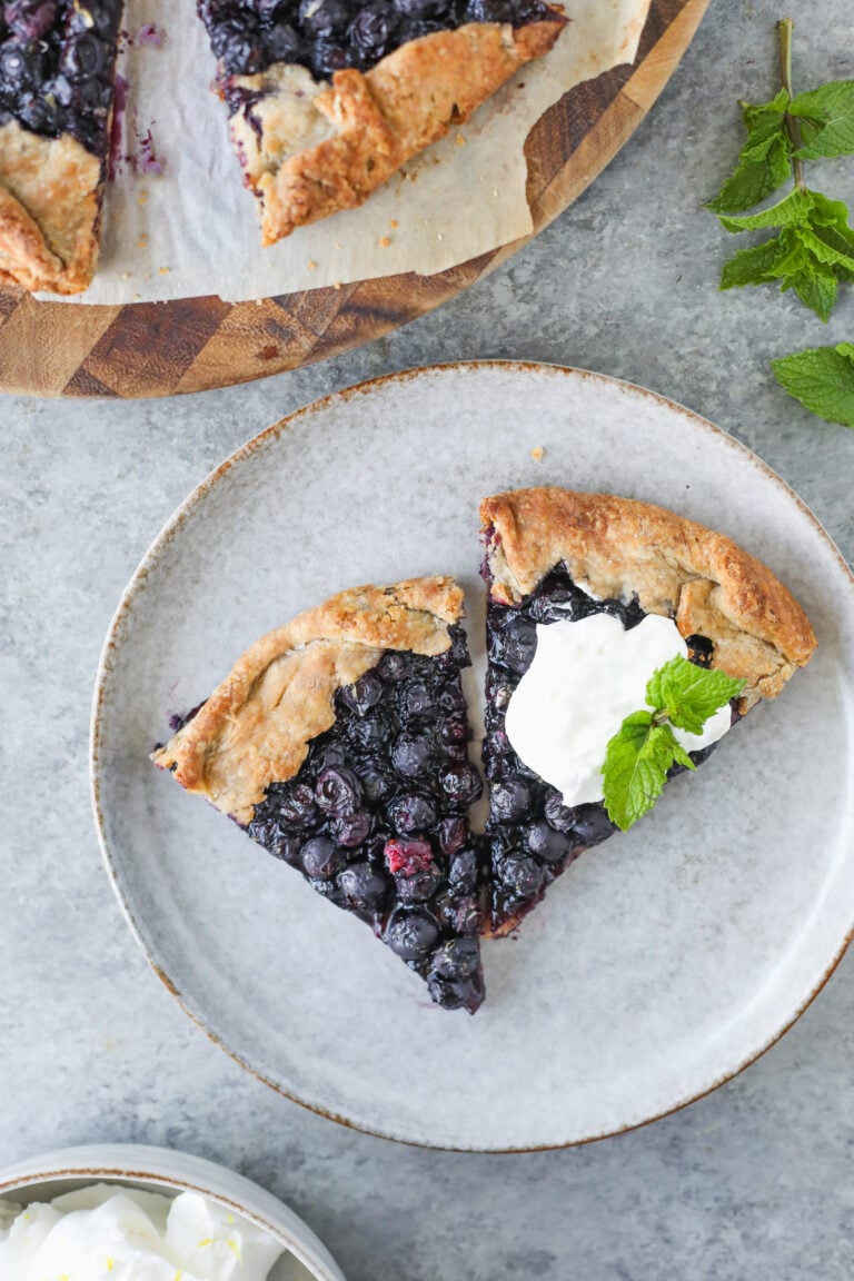 Two Slices Of Blueberry Galette Sit Invitingly On A Plate, One Crowned With A Dollop Of Whipped Cream And A Fresh Mint Sprig. The Plate Rests On A Gray Surface, Surrounded By Extra Blueberry Galette And A Bunch Of Mint Nearby.