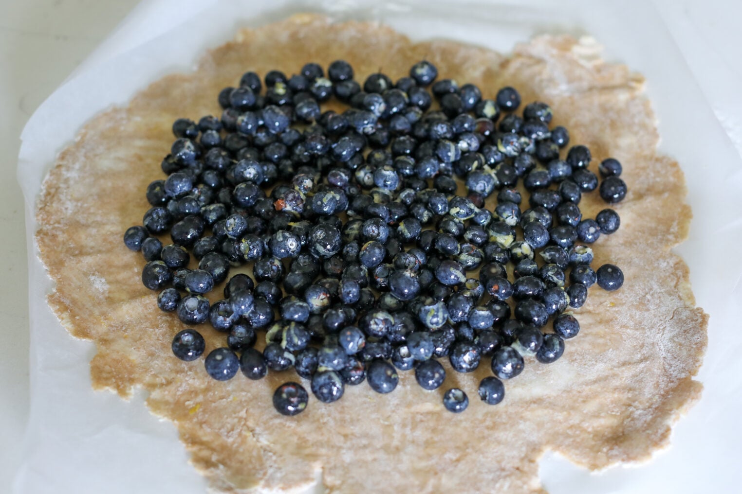 A Pile Of Fresh Blueberries Sits On Rolled-Out Dough Atop A Piece Of Parchment Paper, Ready To Be Made Into A Rustic Blueberry Galette Or Tart.