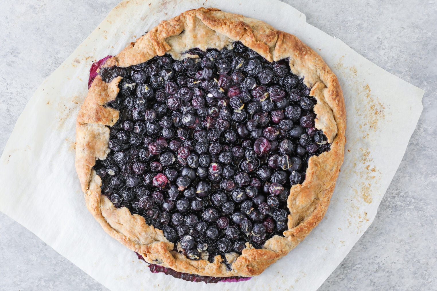 A Rustic Blueberry Galette With A Golden-Brown Crust Sits On Parchment Paper, Displaying Baked, Juicy Blueberries In The Center—The Perfect Embodiment Of A Classic Blueberry Galette.