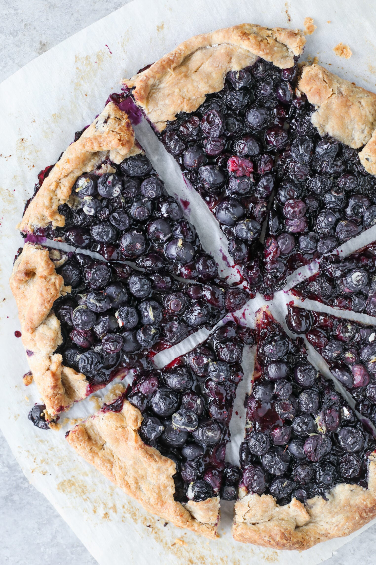 A Rustic Blueberry Galette With A Golden, Flaky Crust, Sliced Into Eight Pieces, Sits On Parchment Paper. Juicy Blueberries Fill The Center Of The Blueberry Galette, With Some Juice Seeping Onto The Crust And Paper.