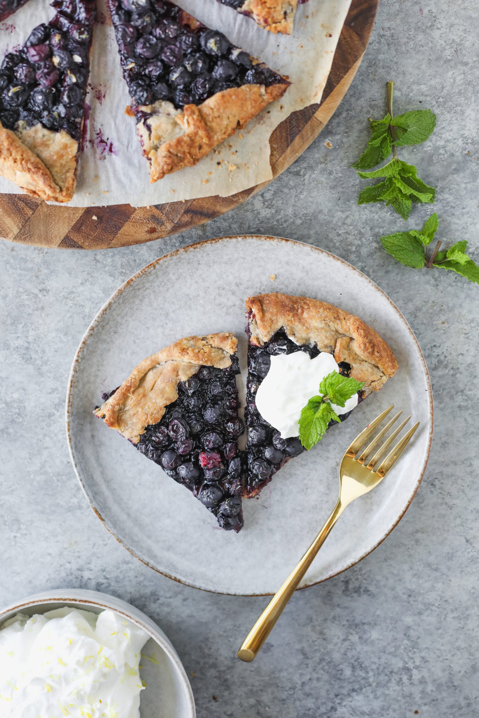 A Plate With Two Slices Of Blueberry Galette, Topped With Whipped Cream And A Mint Leaf, Sits Next To A Gold Fork. A Bowl Of Whipped Cream And More Blueberry Galette Are Nearby On A Gray Surface.