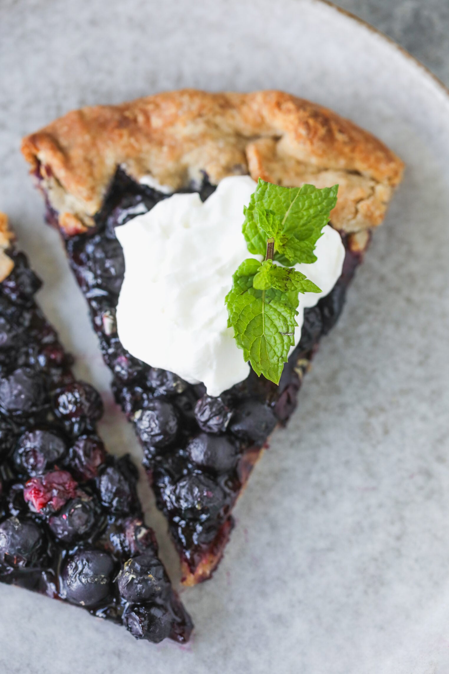 A Slice Of Blueberry Galette Topped With Whipped Cream And A Sprig Of Fresh Mint Sits On A Light Gray Plate.