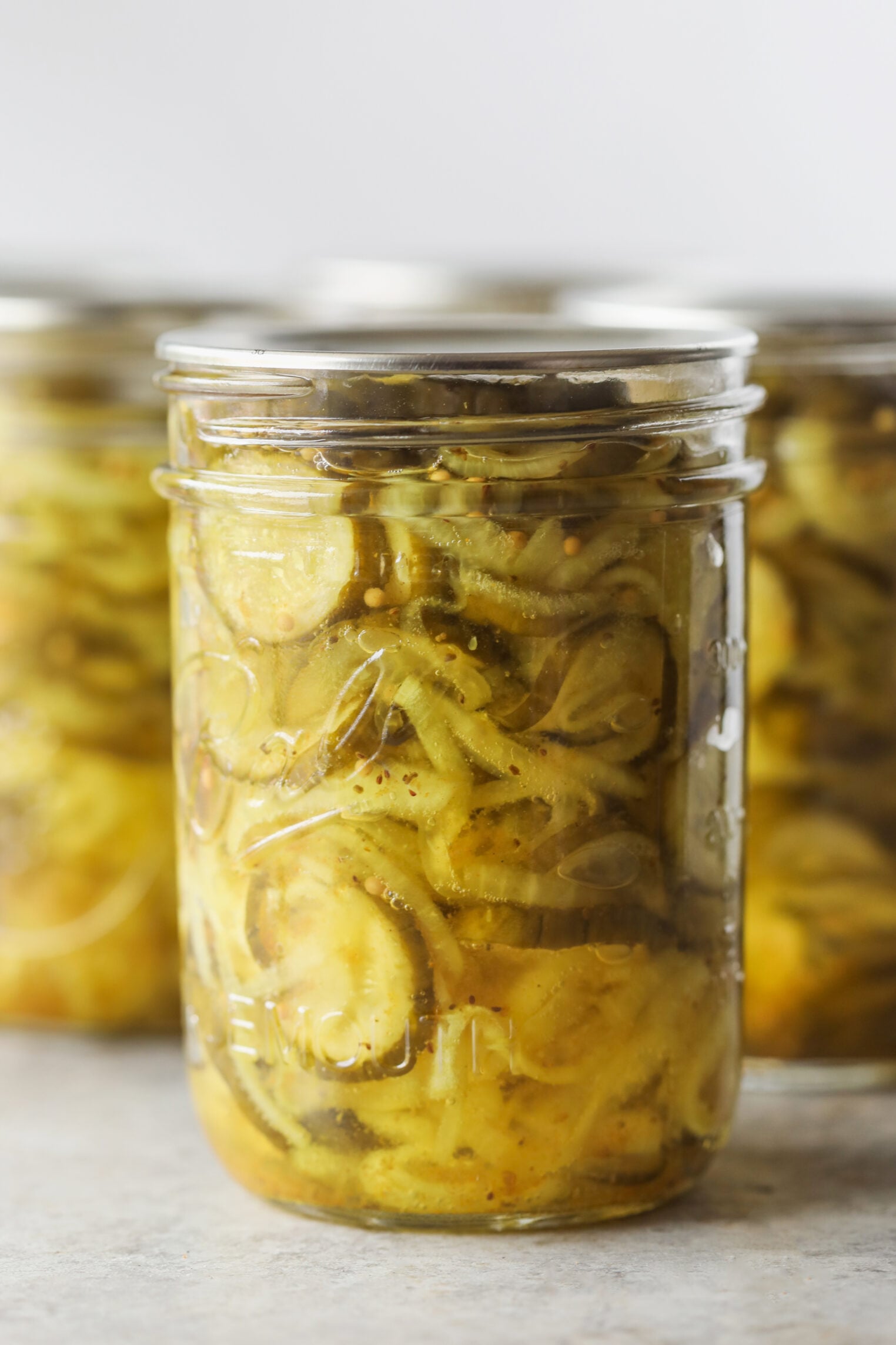 A Close-Up Of A Glass Jar Filled With Bread And Butter Pickles—Crisp Cucumber Slices Immersed In A Yellowish Brine—With Other Similar Jars Blurred In The Background.
