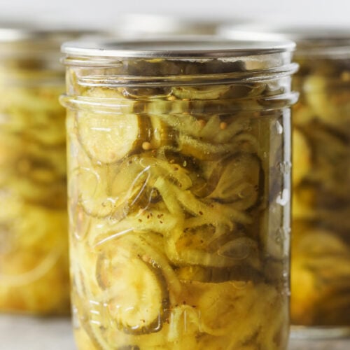 A Close-Up Of A Glass Jar Filled With Bread And Butter Pickles—Crisp Cucumber Slices Immersed In A Yellowish Brine—With Other Similar Jars Blurred In The Background.