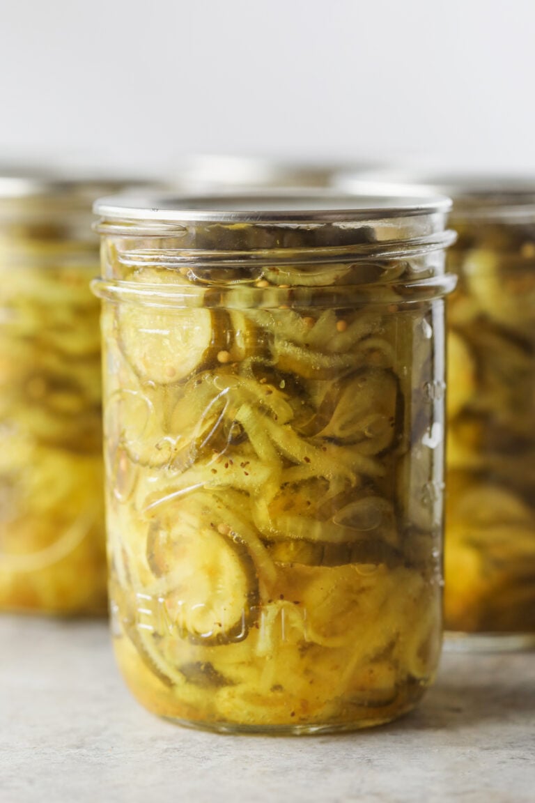 A Close-Up Of A Glass Jar Filled With Bread And Butter Pickles—Crisp Cucumber Slices Immersed In A Yellowish Brine—With Other Similar Jars Blurred In The Background.