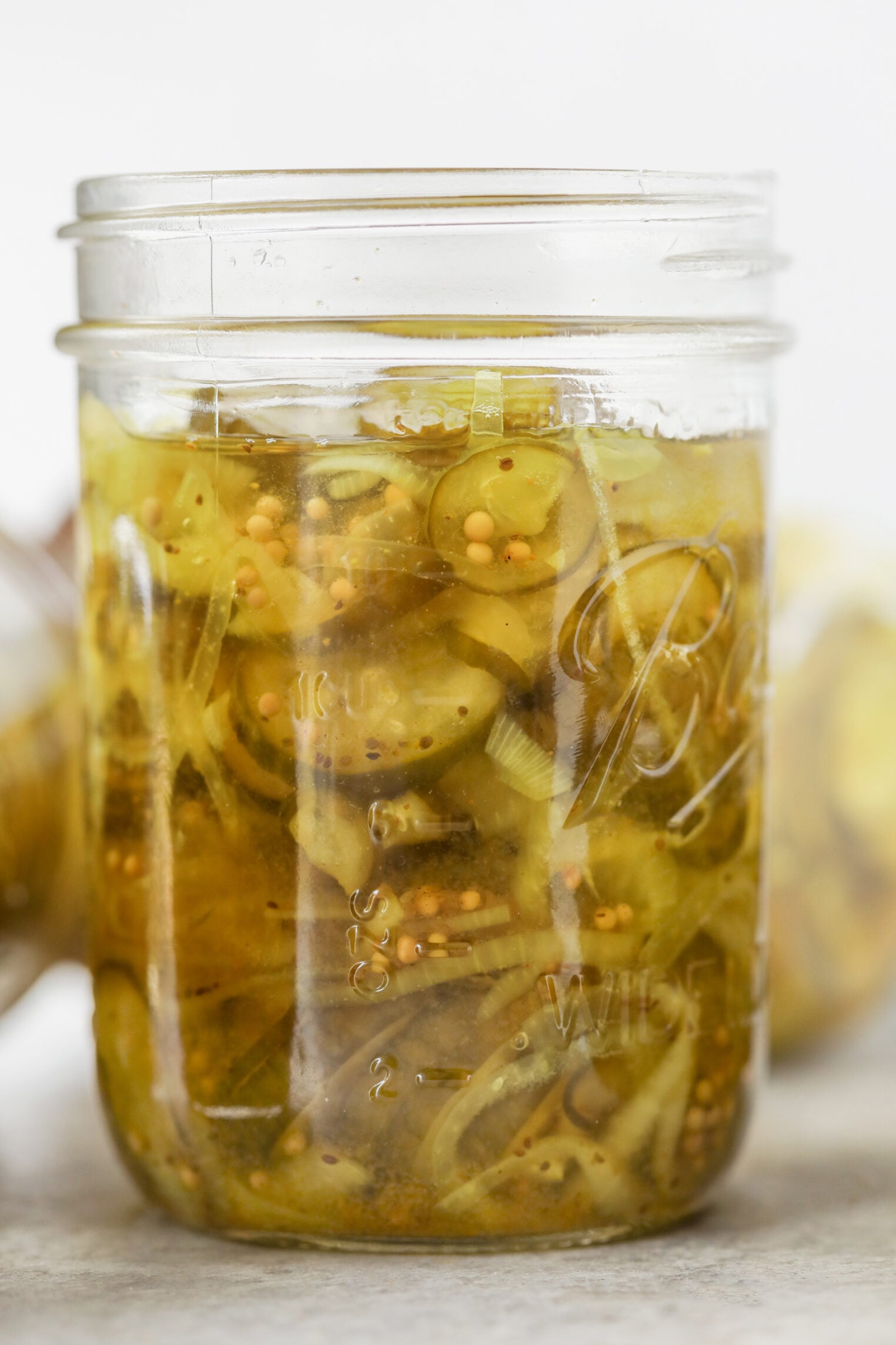 A Glass Jar Filled With Sliced Bread And Butter Pickles And Mustard Seeds In Brine, Sitting On A Light Surface With Another Jar Blurred In The Background.