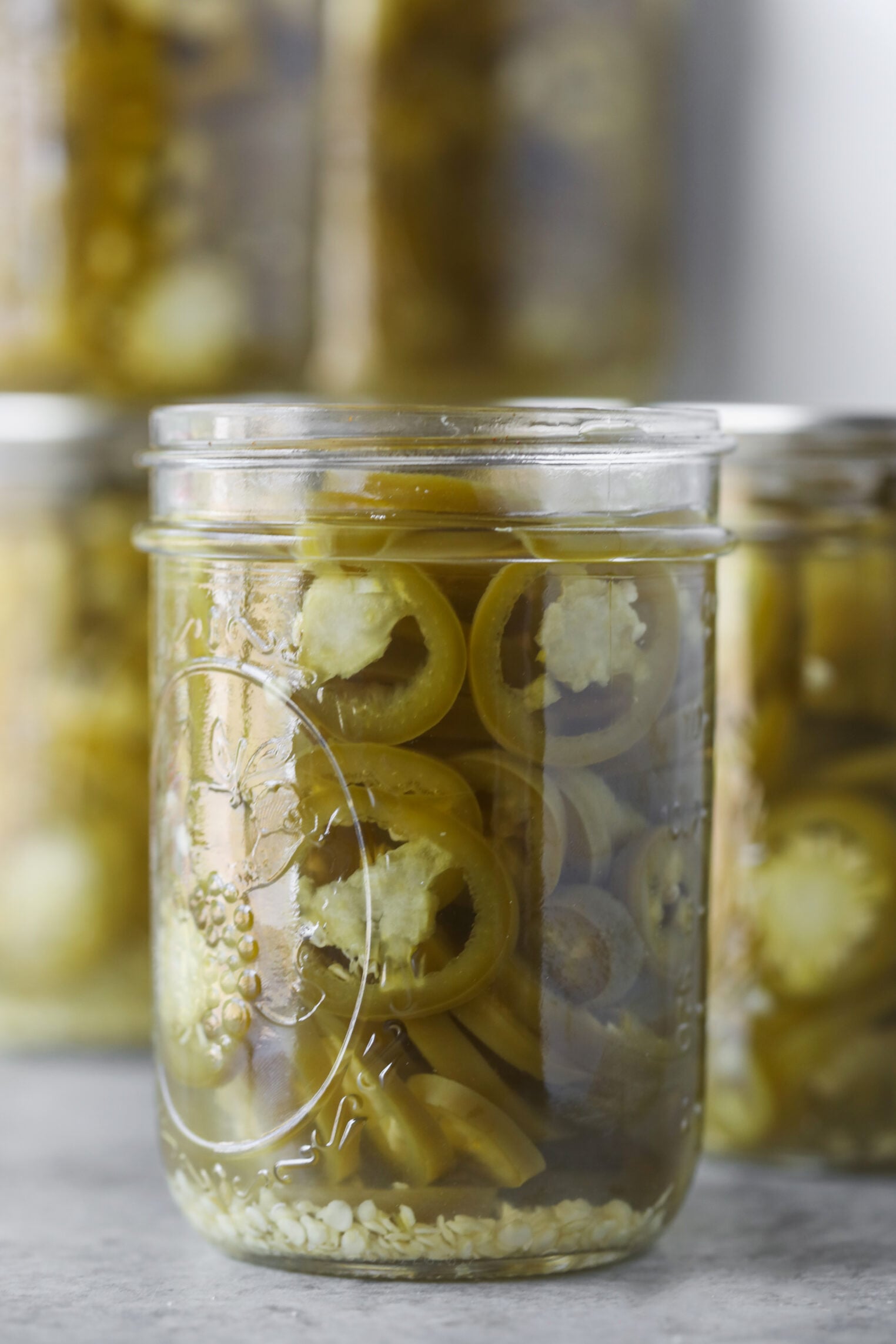 Pickled Jalapeños (Canning Recipe) 10 A Glass Jar Filled With Pickled Jalapeños In Brine, Their Vibrant Slices And Seeds Clearly Visible At The Bottom. Blurred Jars Of Other Pickled Vegetables Can Be Seen In The Background.